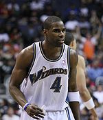 A man of darker complexion wearing a white uniform, on the jersey of which the word "WIZARDS" is printed.