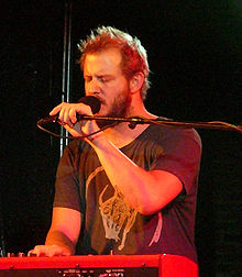 Photographie de Justin Vernon, en train de jouer du synthétiseur d'une main en chantant de l'autre