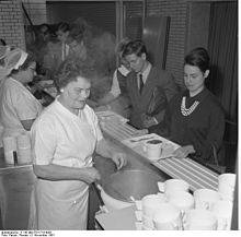 Photographie en noir et blanc. Des femmes en uniforme blanc servent les portions de nourriture sur les plateaux qu’étudiants et étudiantes font glisser devant elles sur un comptoir rainuré. Les étudiants sont en costume-cravate, les étudiantes sont élégamment vêtues.