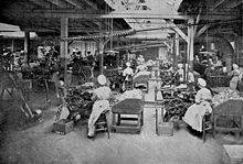 Photographie en noir et blanc d’une immense salle que les verrières du plafond, qui repose sur des poutres et des pilastres, éclairent chichement. À perte de vue se répètent les rangées de mêmes machines actionnées par des femmes assises, en coiffes et longs tabliers blanc. De rares hommes, déambulent dans les allées, surveillant…