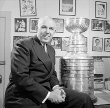 Photo noir et blanc de Clarence Campbell qui pose assis sur le coin d'un bureau à côté de la Coupe Stanley et devant un mur couvert de photos.