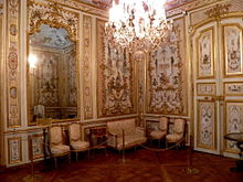 Vue de deux murs d'un cabinet décoré de boiseries blanches et de dorures, couvertes de peintures, avec quatre chaises et un canapé