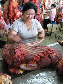 Une femme typiquement asiatique découpe un gros quartier de carcasse sanguinolente le long es côtes à l'aide d'un long couteau, la rapidité de l'action étant suggérée par le flou de la photo.