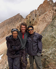 Trois hommes sont debout en extérieur, face à l'appareil photo. Au fond, les sommets de montagnes. Le plus grand, un Occidental barbu, se trouve au milieu, ses bras sur les épaules de deux Asiatiques plus petits.