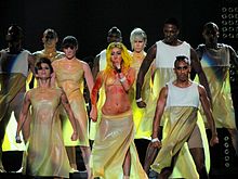 Gaga portant une perruque jaune et une tunique de couleur chaire accompagnée de ses danseurs tous debouts sur scène.