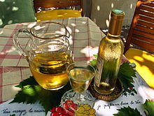 Photographie en couleur illustrant bien la chanson « Ah, le petit vin blanc... ». Une carafe de vin blanc avec un verre plein et une bouteille bouchée sont posés sur des feuilles de vigne, elles-même sur une nappe blanche à carreaux rouges. La bouteille couverte de buée donne une sensation de fraîcheur et le jeu d'ombre et de lumière laisse penser que la scène est située sous une tonnelle,