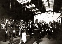 Suivi par une cohorte d’officiels, la reine, entourée par le roi Albert et le roi Fouad, avance, portant un bouquet de fleurs, sur le tapis déroulé sous l’auvent d’une gare.
