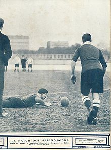 Un joueur de dos se prépare à taper dans le ballon posé au sol devant un autre joueur à plat ventre sur le sol