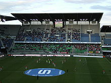 Photographie montrant une vue d'ensemble de la tribune Rennes et de ses spectateurs, avec un écran géant dans sa partie haute et la partie réservée aux supporters adverses sur son côté droit.