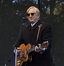 Photographie montrant T-Bone Burnett sur scène, jouant de la guitare à la main, devant un micro, habillé de noir et portant des lunettes noires.