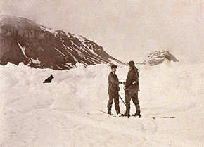  Deux hommes se serrent la main au milieu d'un champ de neige, un chien à leurs côtés. On aperçoit de sombres collines à l'arrière-plan.