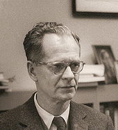 Photographie en noir et lanc du psychologue américain Burrhus Frederic Skinner, auteur de « Walden Two » (1948). L'avant du crâne légèrement dégarni et portant des lunettes, B. F. Skinner est habillé d'un costume et d'une cravate. Derrière lui, une étagère de livres.