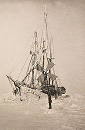  Vue de face légèrement décalée d'un navire couvert de givre, entouré de monceaux de glace. Un personnage solitaire se trouve debout sur la glace à côté du navire.