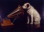 Un chien blanc à droite regarde dans l’œil du cône d’amplification d’un gramophone à gauche.