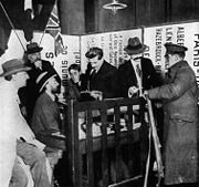 Photo en noir et blanc. Derrière une table-bureau abritée par une barrière en bois se pressent six hommes affairés en complet veston, pour la plupart chapeautés ; devant la barrière un soldat anglais reçoit un ticket de repas (il « est vu acceptant un ticket » dit la légende en anglais. La scène se passe dans un angle réduit formé de panneaux d’avis et donne une impression de confusion.