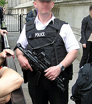 Agent de police en uniforme portant un gilet pare-balles réglementaire et un fusil d'assaut, dans une rue.