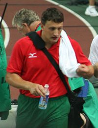 Un homme habillé en rouge et en vert tient une bouteille d'eau de la main gauche et une serviette de la main droite.