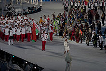 Un athlète en blanc et rouge tient le drapeau biélorusse puis un groupe d'athlètes biélorusses est derrière lui.