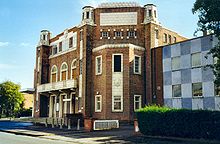 A large brick building. A more modern extension goes off to the right. The building sits on the corner of a street, and is seen in bright, sunny weather.