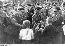 Photographie montrant deux enfants, une fille à gauche et un garçon à droite remettant un bouquet de fleurs à Heinrich Himmler au centre de la photographie ; le personnage de gauche est Wilhelm Koppe, celui de droite Fritz Bracht.