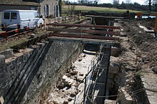  Photo couleurs. L’écluse du bas de Grignon, vue en perspective frontale, est en travaux. Elle est à sec, on peut voir au fond de la photo, les palplanches faisant office d’étanchéité. A droite, le mur de droite de l’écluse (bajoyer) est entièrement démonté pour que l’ensemble des pierres le constituant soit remplacé par des pierres de taille neuves. Sur la partie gauche, seul le couronnement (partie haute du mur) est démonté. Une passerelle métallique de chantier traverse l’écluse. Des pierres de taille neuves reposent ne fond d’écluse.