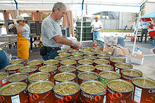 Sous un chapiteau, une quarantaine de grandes boites de haricots verts en morceaux sont ouvertes et prêtes à être cuisinées ; deux personnes, gantées, s’en occupent.