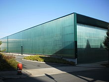 Bâtiment rectiligne aux parois de verre teinté, dans lesquelles se reflète le ciel bleu.