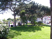 Photographie du parc montrant des allées passantes, des pelouses, des massifs de fleurs et des plantations exotiques à l'ombre des pins parasols. Au fond à droite, la courbe des bâtiments à galeries et loggias du front de mer.