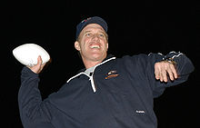 Photographie de John Elway en survêtement et coiffé d'une casquette qui pose de nuit avec un ballon en main
