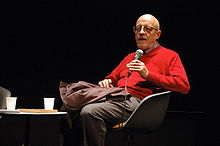 Le cinéaste Luc Moullet en 2008 à la cinémathèque française, il est vêtu d'un pull rouge et d'un pantalon gris. Il parle dans un micro pour se faire entendre du public.