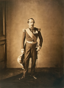 Photographie en sépia de Napoléon trois, de plein pied. Il port un uniforme militaire, et tient un bicorne de sa main droite. Son visage arbore un air éteint, comme sur de nombreuses photographies.