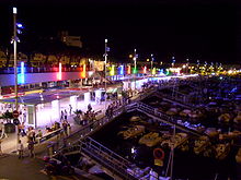 Photographie présentant un panorama du port la nuit: au premier plan des pontons de bois et des bateaux blancs, au second plan les quais illuminés où circulent des promeneurs.