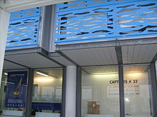 Photographie d'un bâtiment à la structure de métal galvanisé et lattes blanches, formant une galerie abritant des vitrines. À l'étage, des balcons bleu vif ajourés de formes ondulées évoquant des vagues