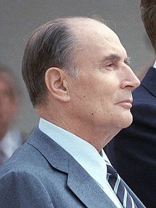 Photographie de François Mitterrand, en 1984, avec un gros plan sur le haut du buste et sur sa tête. Il est pris presque de profil, depuis la droite. Son air est solennel.