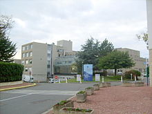 Photographie de l'hôpital montrant des bâtiments parallélépipédiques imbriqués avec au premier plan le volume vitré de l'entrée principale, ombré par des claustras horizontales et quelques beaux arbres.