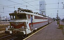 La photographie couleur montre une locomotive CC 40100, couleur inox et rouge, attelée à un train de voiture de même couleur., à quai en gare de Bruxelles-Midi.